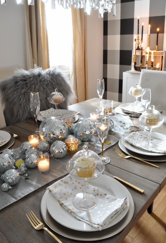 a shiny holiday tablescape with silver disco balls forming a table runner, candles, white porcelain and gold cutlery