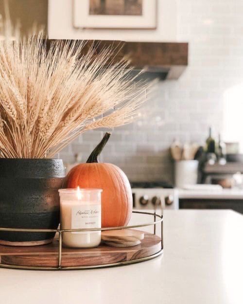 a simple fall or Thanksgiving decoration of a wooden tray with a pumpkin spice candle, a pumpkin and some wheat in a planter