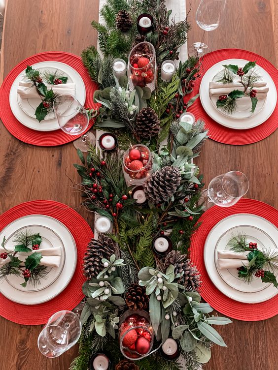 a stylish Christmas tablescape with evergreens, pinecones, candles, red placemats, white porcelain and napkins accented with greenery
