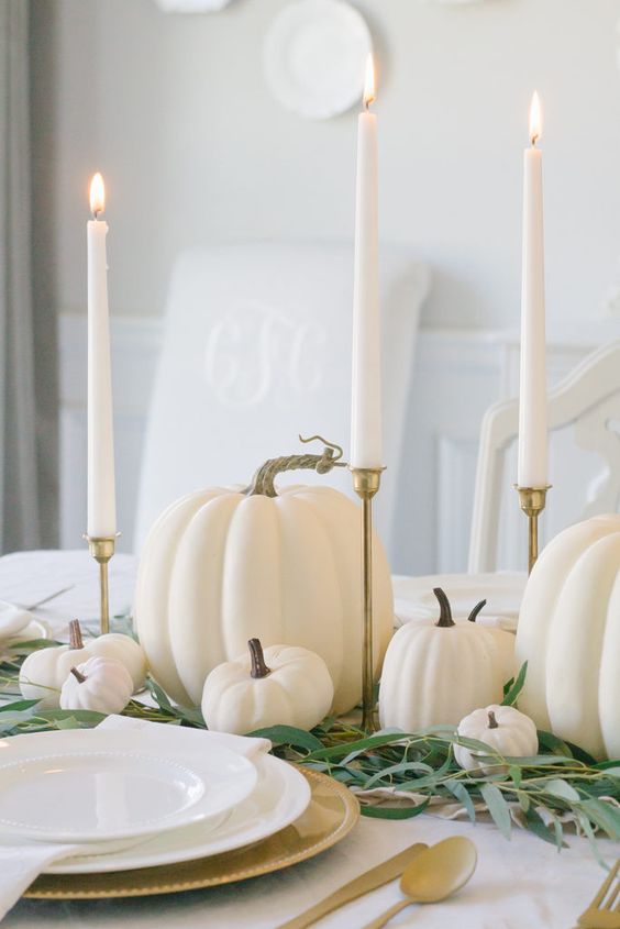 a super easy and cool Thanksgiving centerpiece of greenery, white pumpkins, tall and thin candles in gold candleholders is lovely