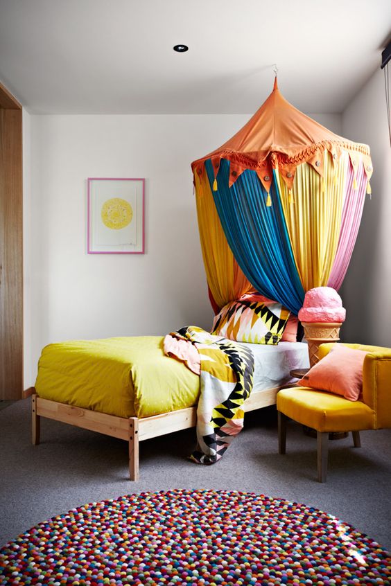 a unique kid's room with a bed with a colorful circus-inspired canopy and bedding, a yellow chair and a colorful pompom rug
