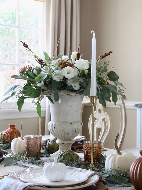 a vintage rustic Thanksgiving centerpiece of an urn with white and rust blooms, greenery and much texture plus a tall and thin candle