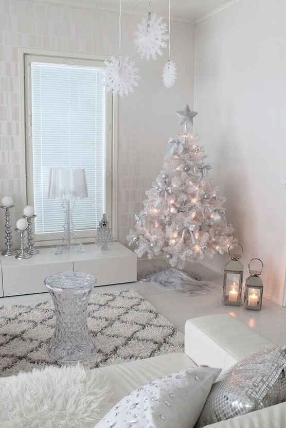 a white and silver living room with a white Christmas tree with lights and silver ornaments, white snowflakes hanging from the ceiling and shiny pillows