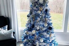 a blue Christmas tree with white, blue, navy, gold and silver ornaments, branches with pompoms and white ribbons for a beach or coastal space