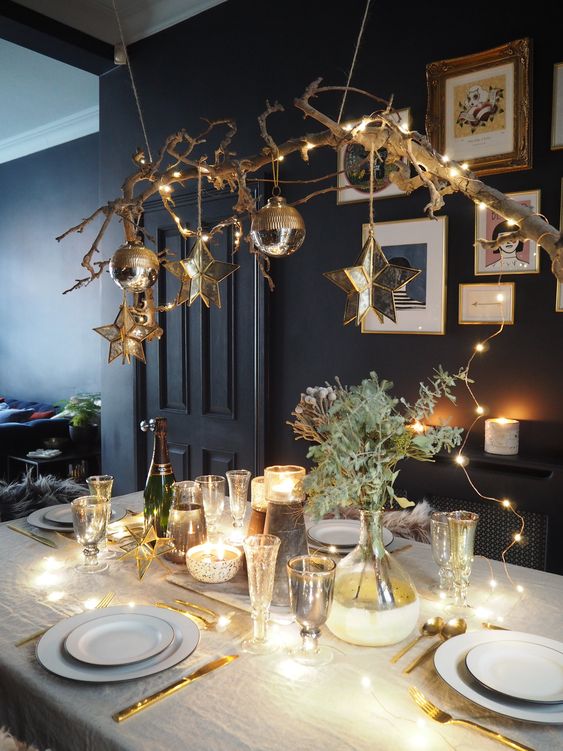 a chic Christmas tablescape with a branch with ornaments and stars hanging over the table, lights and greenery on the table and gold cutlery