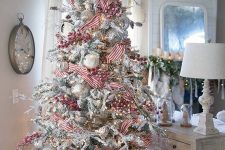 a flocked Christmas tree with oversized white and silver ornaments, red berries and striped ribbons plus lights