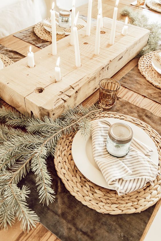 a hygge Christmas table with woven placemats, evergreens, striped napkins, a wooden board with tall and thin candles