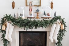 a lovely Christmas mantel with a lush evergreen garland, cloches with mini trees and gift boxes, oversized balls and stockings