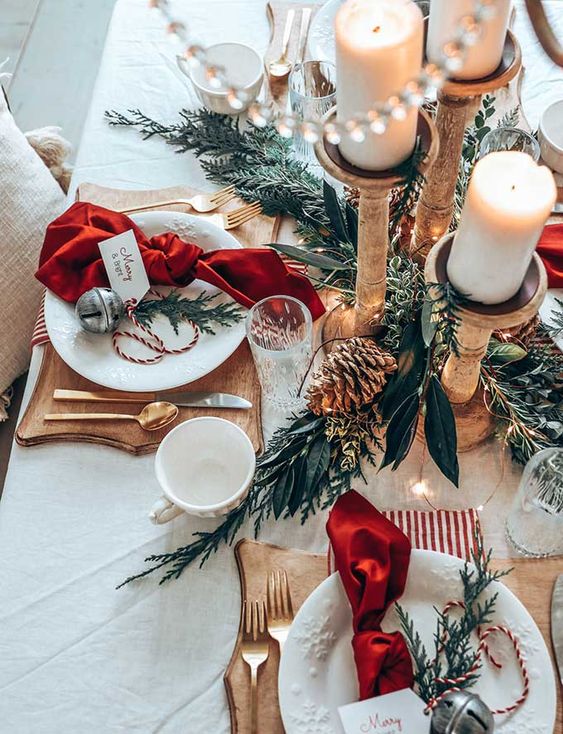 a lovely Christmas tablescape with greenery, pinecones, wooden candleholders with pillar candles, red napkins, cutout wooden boards and bells