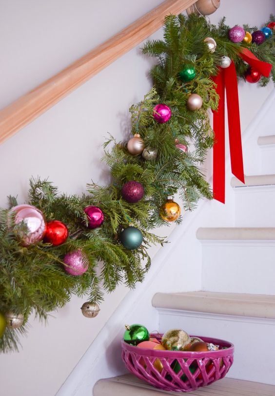 a lush evergreen and colorful Christmas ornament garland with red bows is a cool railing decor idea