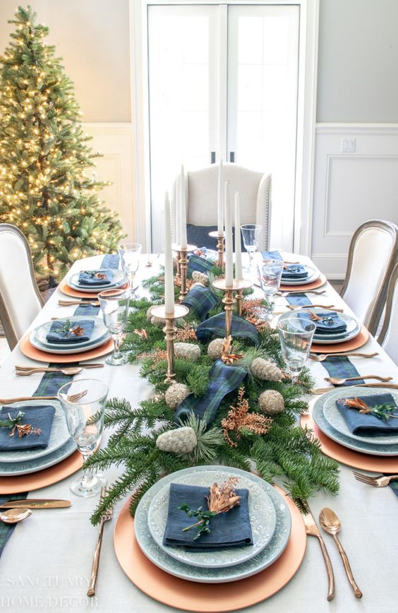 a pretty Christmas tablescape done in navy and copper, with tall and thin candles, printed plates, an evergreen and pinecone runner plus a plaid ribbon