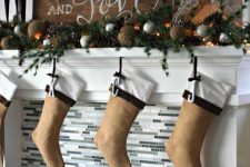 a rustic Christmas mantel with an evergreen, pinecone and silver ornament garland, candles, lights, a wooden sign and burlap stockings