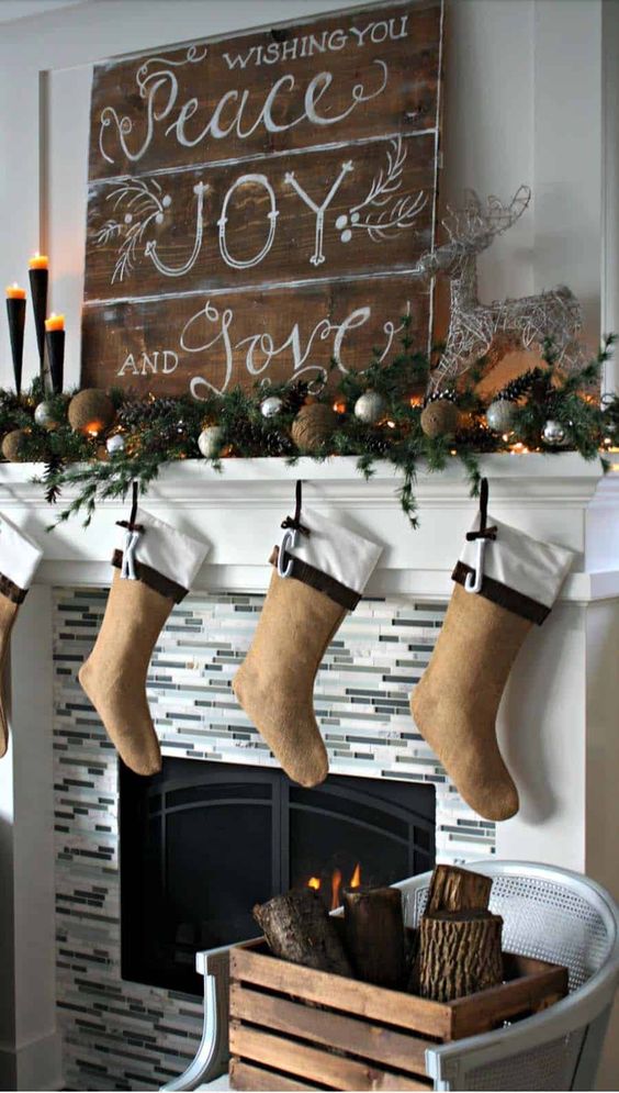 a rustic Christmas mantel with an evergreen, pinecone and silver ornament garland, candles, lights, a wooden sign and burlap stockings