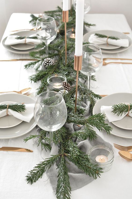 a simple modern Christmas tablescape with an evergreen runner, snowy pinecones, tall and thin candles, neutral porcelain and linens