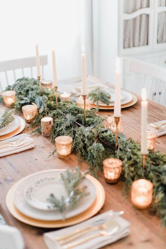 an elegant Christmas table setting with an evergreen runner, tall and thin candles and small candleholders, gold chargers and printed plates