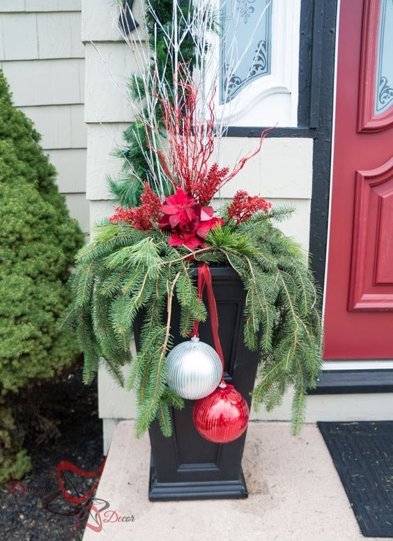 an outdoor urn with evergreens, white branches, red blooms and a giant silver and red ornament