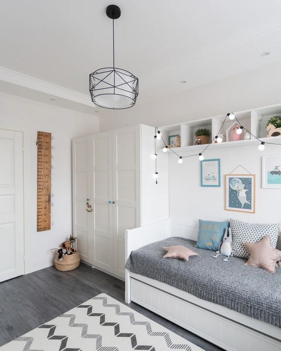 a Nordic kid's room with a wardrobe, a bed with grey bedding, box-shaped shelves, string lights and a printed rug