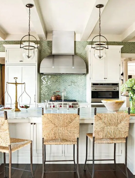 a beautiful coastal kitchen with white vintage cabinets, a large kitchen island with wicker chairs, glass pendant lamps and a green tile backsplash