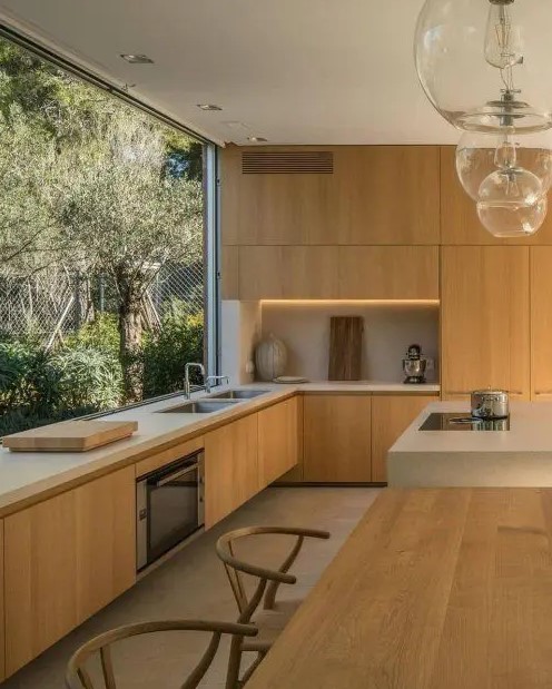 a beautiful contemporary kitchen with sleek light-stained cabinets, white stone countertops, built-in lights and a glazed wall with garden views