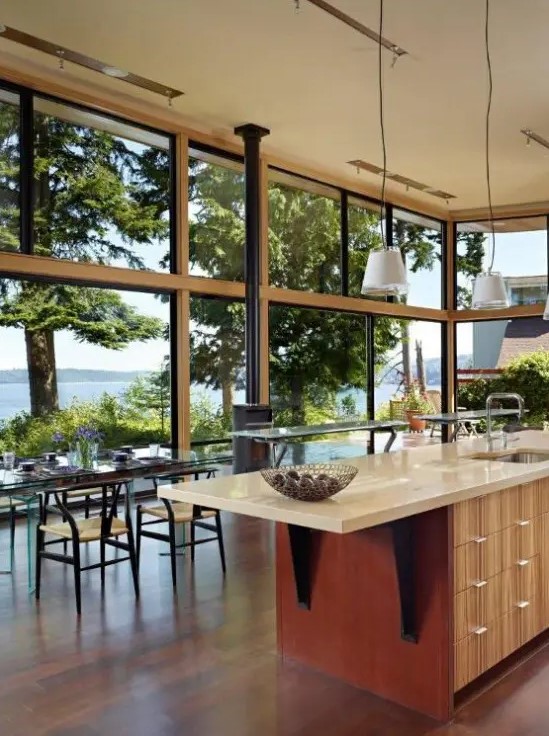 a beautiful modern kitchen and dining space in one, with glazed walls, a hearth, a light-stained kitchen island, a glass table and woven chairs is chic