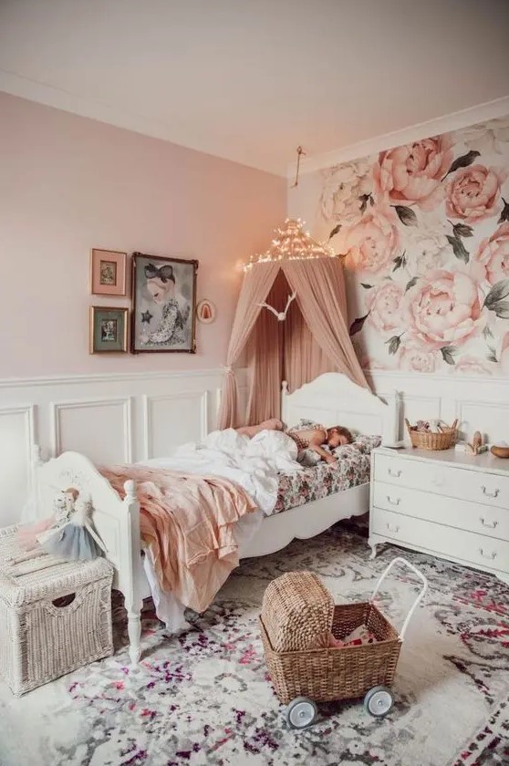 a beautiful pink girl's bedroom with floral wallpaper, white paneling, vintage white furniture, a printed rug and a dusty pink canopy over the bed