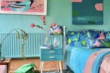 a blue and green bedroom with turquoise walls, a blue nightstand, a bed with pink and blue bedding and lovely artworks