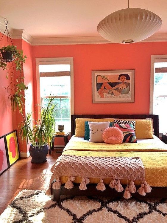 a bold bedroom with coral walls, a dark bed with colorful bedding, a tassel blanket, colorful artworks and potted plants