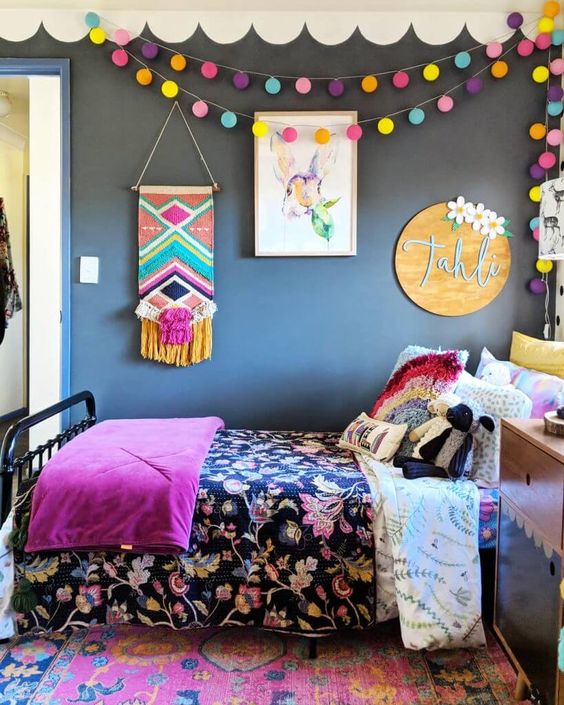 a bold teen bedroom with graphite grey walls, a black bed with colorful bedding and pillows, a bright bunting and a macrame hanging and a bold printed rug