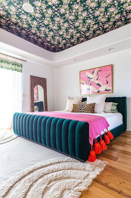 a bright bedroom with a floral ceiling, a teal upholstered bed, bright bedding, a colorful artwork and a mirror in a carved frame