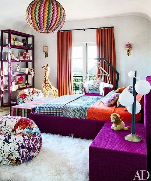 a bright bedroom with a purple bed and nightstnads, a purple storage unit, a floral pouf, orange curtains and a colorful pendant lamp