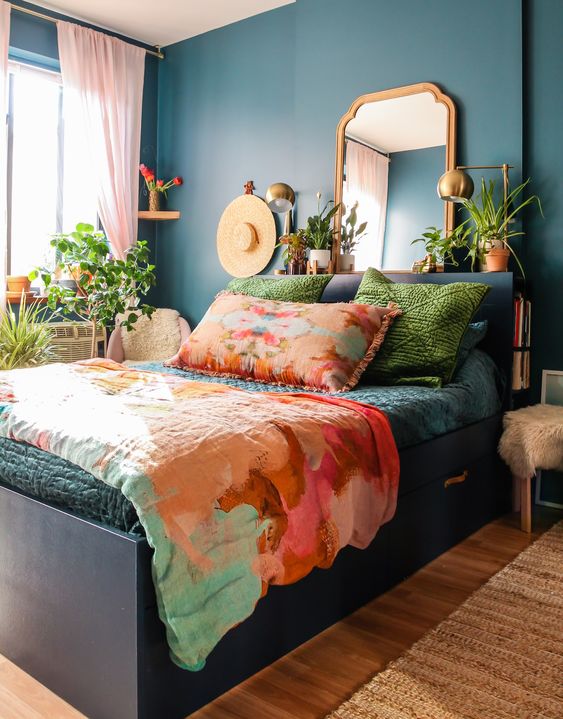a bright bedroom with navy walls and a black storage bed, colorful bedding, potted plants, a mirror and brass lamps is a fun and bold space
