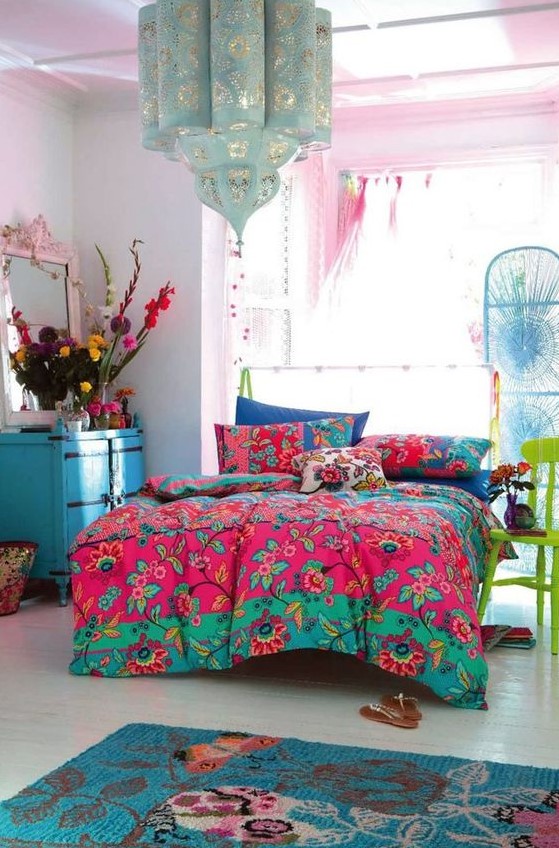a colorful eclectic bedroom with pink walls, a bed, a blue chest of drawers, a green chair, colorful bedding and bold blooms plus a lovely pendant lamp