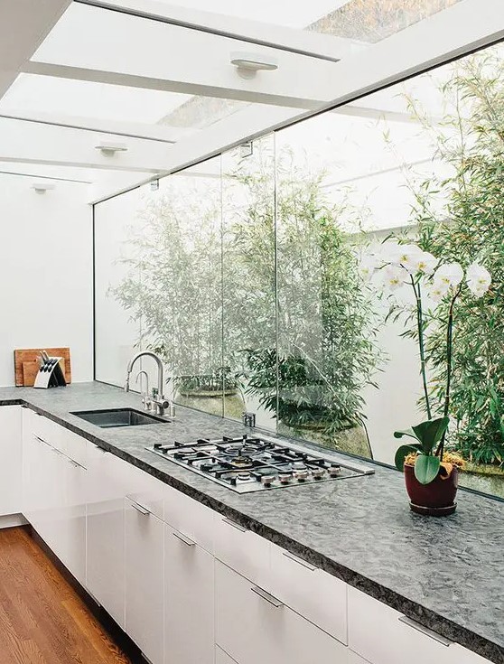 a contemporary white kitchen with a dark countertop, a glazed wall with a view of the private garden and greenery