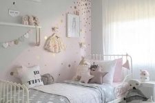 a delicate Scandinavian girl’s room with ledges with artwork and toys, a white bed with mauve and grey bedding, a color block wall with polka dots