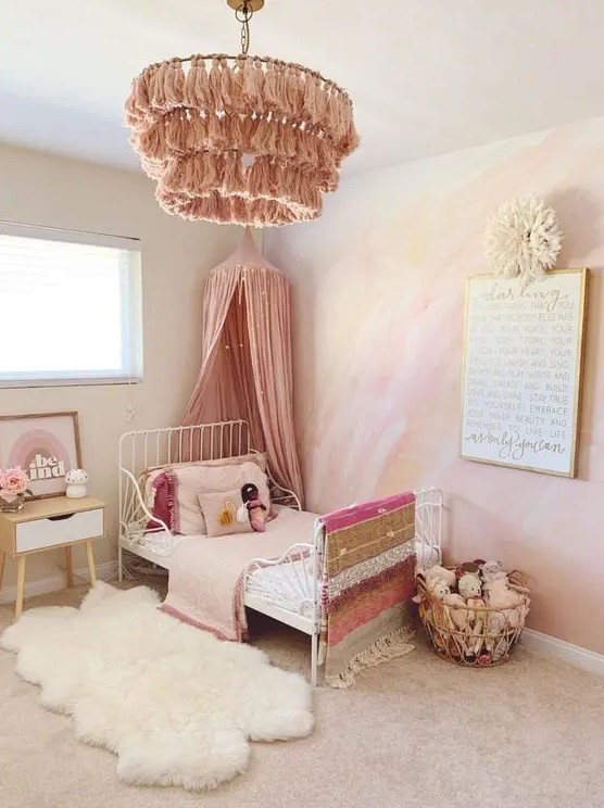 a dreamy pink girl's bedroom with a watercolor yellow and hot pink accent wall, a white metal bed with pink bedding, a pink tassel chandelier