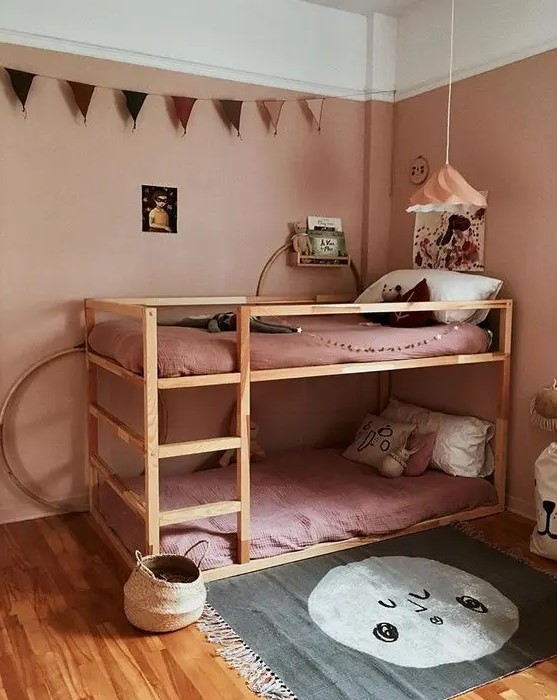 a dusty pink kid's room with dusty pink walls and bedding, an IKEA Kura bunk bed, a printed rug, a garland and baskets