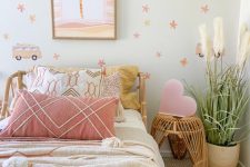 a fun girl’s room with printed wallpaper, a rattan bed with pastel bedding, a rattan nightstand with a pink heart, a potted plant and tassels
