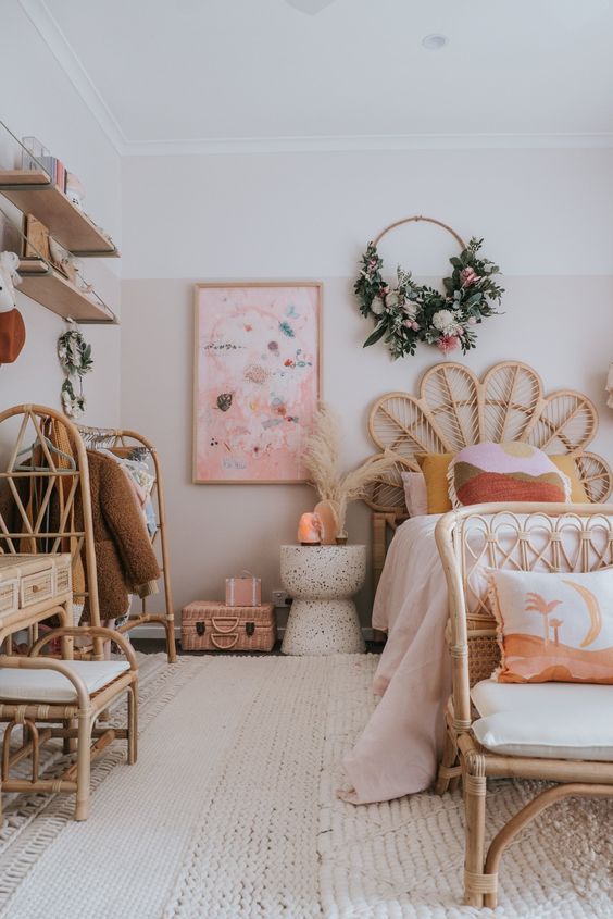 a gorgeous pink boho girl's room with a light pink clor block, rattan furniture with neutral upholstery, wall-mounted shelves and blooms
