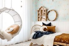 a lovely celestial girl’s room with air balloon wallpaper, a rattan bed with rust and navy bedding, a pendant chair with a pillow and star garlands