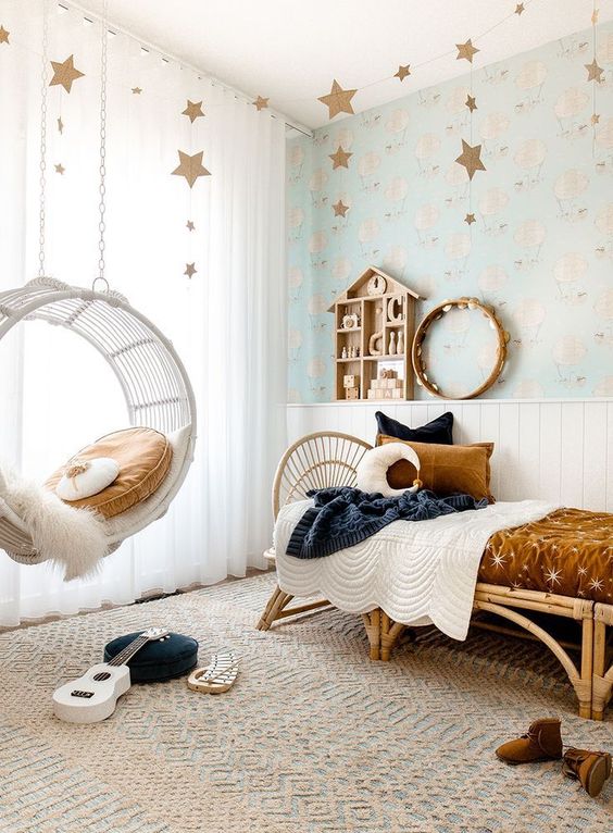 a lovely celestial girl's room with air balloon wallpaper, a rattan bed with rust and navy bedding, a pendant chair with a pillow and star garlands