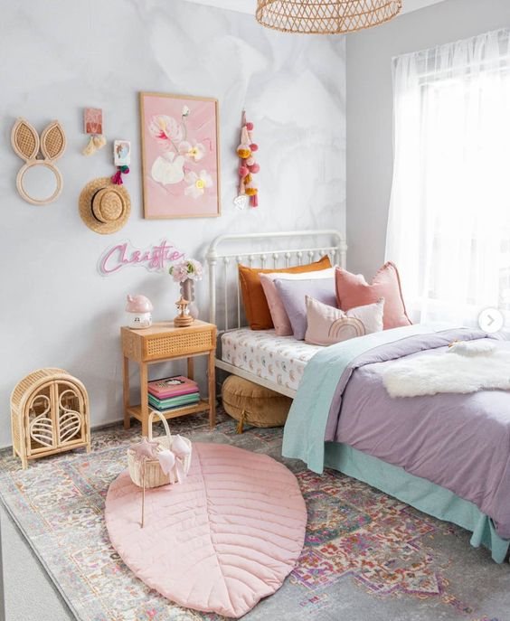 a lovely girl's room with a metal bed with pastel bedding and layered rugs, a rattan nightstand and a storage unit, a gallery wall