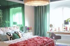 a maximalist bedroom with simple furniture, green mirrors and curtains, a colorful rug and bold bedding plus a fluffy chandelier