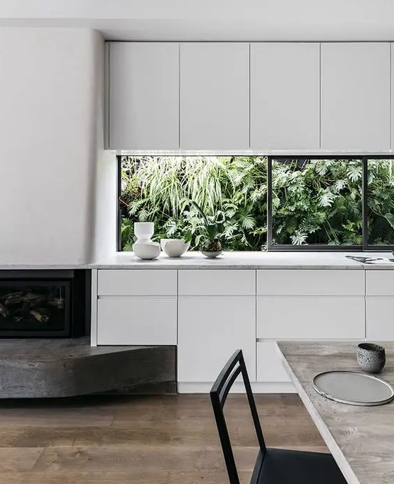 a minimalist white kitchen with sleek cabinets, white stone countertops, a window backsplash that opens on the tropical garden