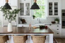 a modern country kitchen in white, with cool cabinets, a large kitchen island with a butcherblock countertop, pendant lamps and woven stools