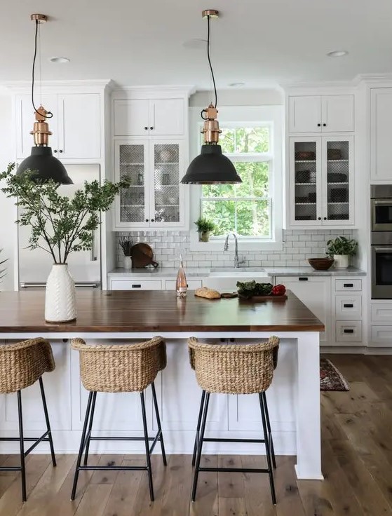 a modern country kitchen in white, with cool cabinets, a large kitchen island with a butcherblock countertop, pendant lamps and woven stools