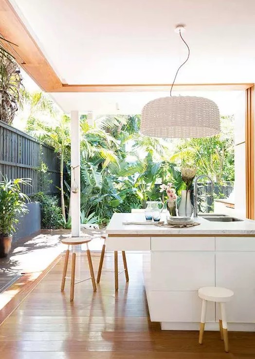 a modern white kitchen with a large woven lampshade and white stools, with walls that can be removed to connect to the tropical garden