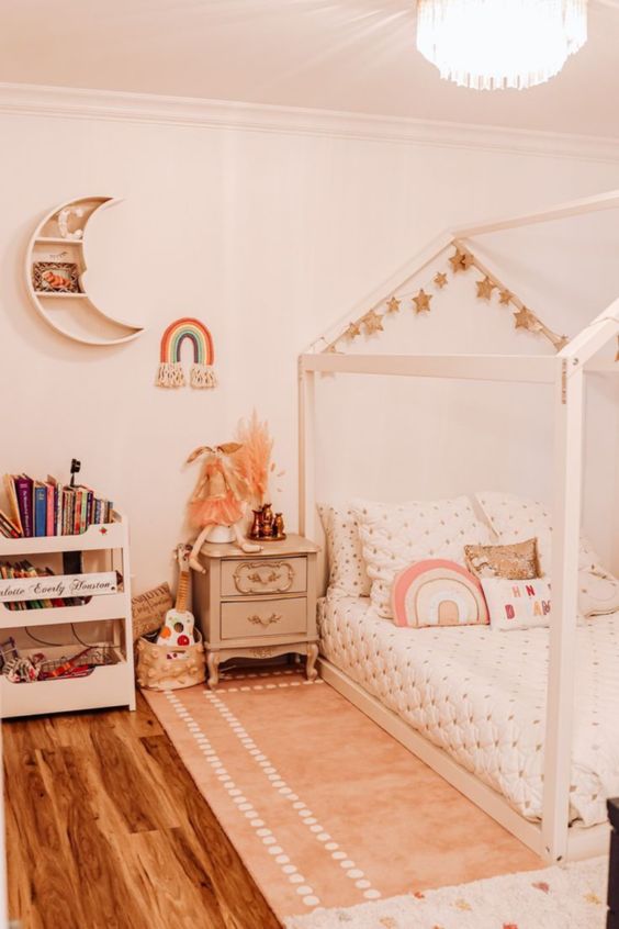 a neutral boho girl bedroom with a wooden house-shaped bed with neutral bedding, a storage unit, a vintage nightstand and a moon-shaped shelf