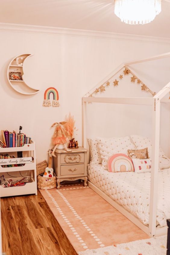 a neutral boho girl bedroom with a wooden house-shaped bed with neutral bedding, a storage unit, a vintage nightstand and a moon-shaped shelf