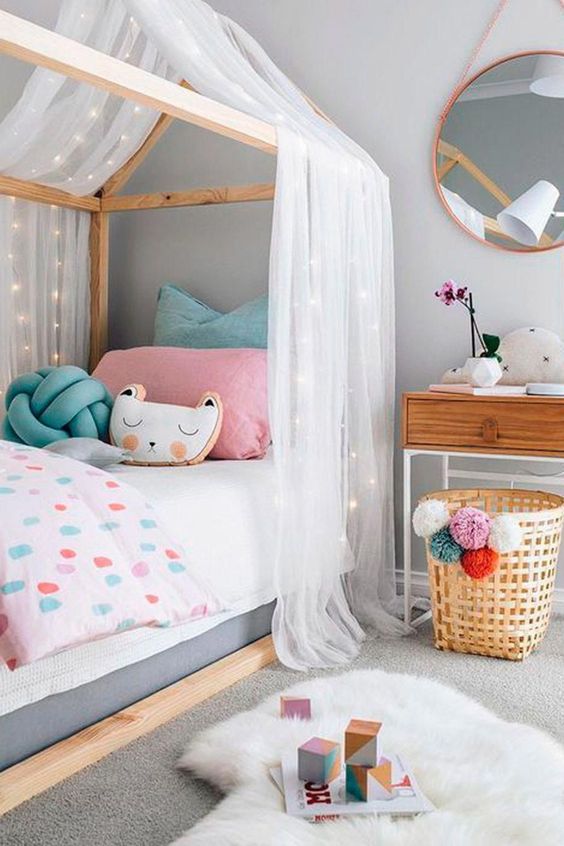 a neutral girl's room with a house-shaped bed with pastel bedding, a nightstand, a basket with pompoms and layered rugs