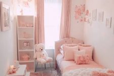 a pink and white girl’s bedroom with an upholstered bed with pink bedding, pink curtains, a fluffy lamp, a white shelving unit and gallery walls